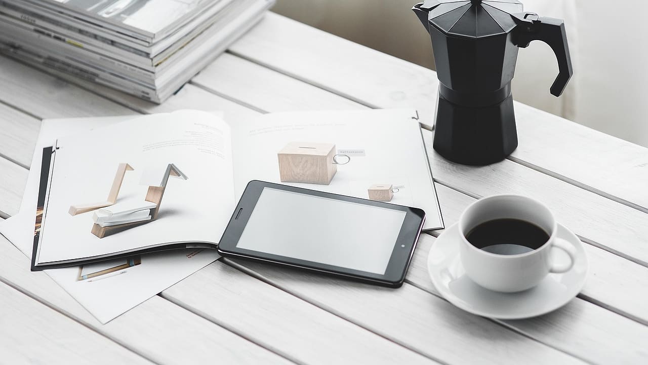 Tablet, filiżanka kawy i kawiarka na białym biurku obok stosu magazynów