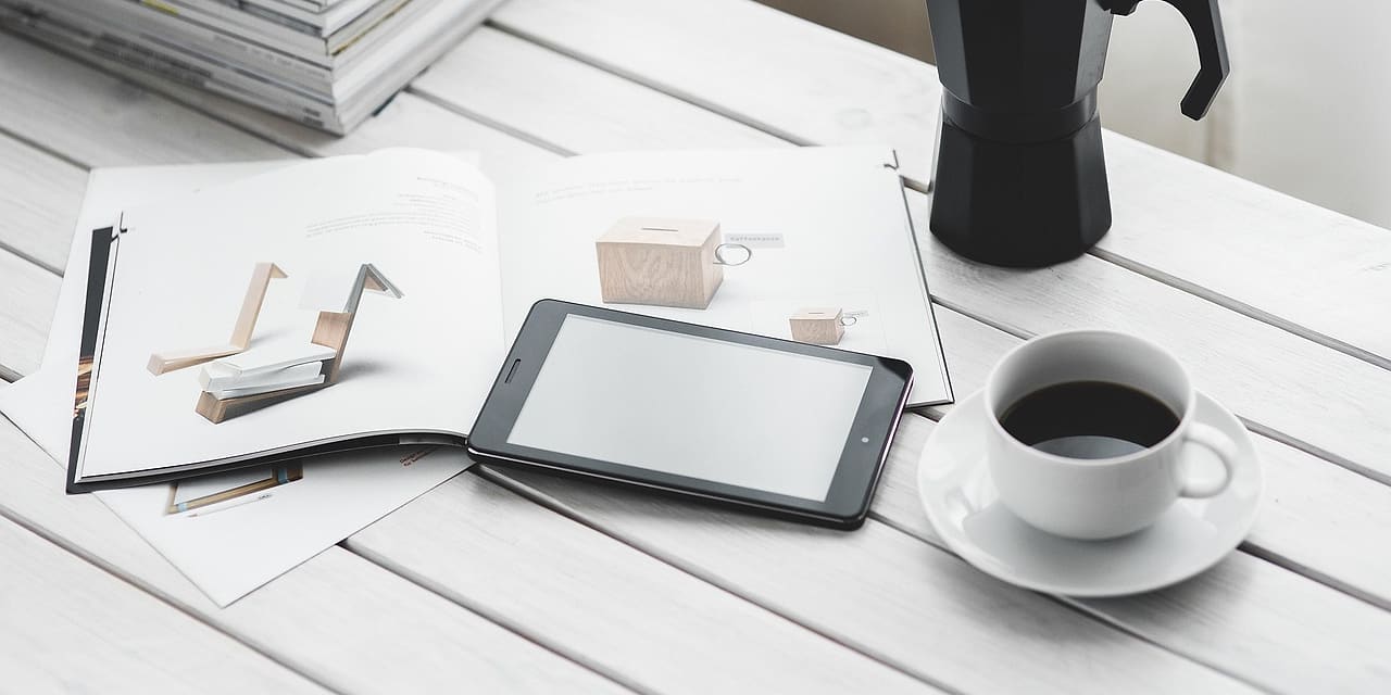 Tablet, filiżanka kawy i kawiarka na białym biurku obok stosu magazynów