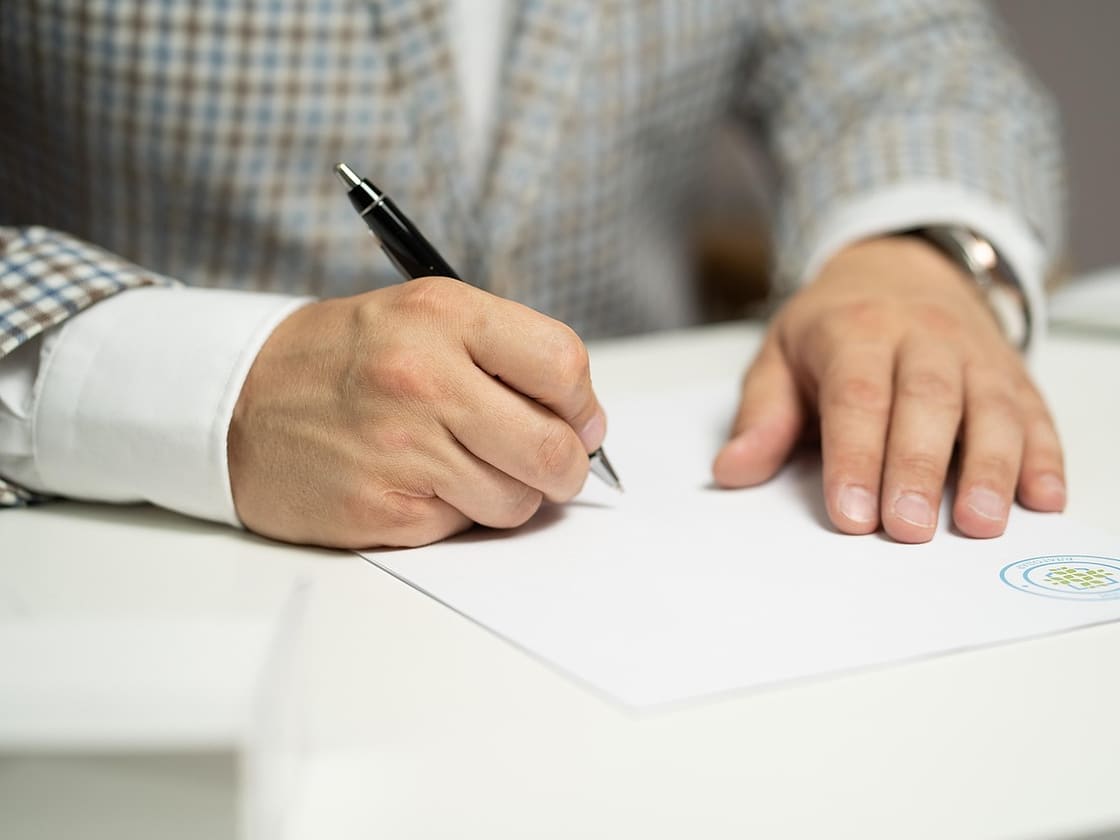 Mężczyzna w marynarce podpisuje dokument piórem przy białym biurku