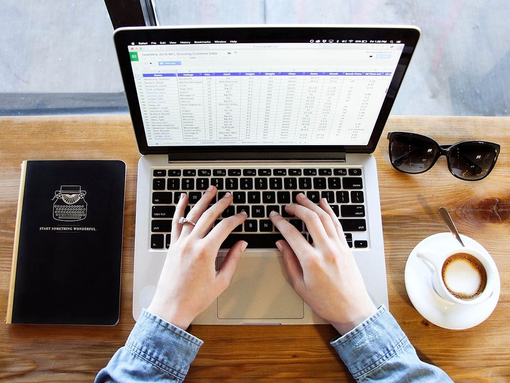 Ręce wpisują dane w arkusz na laptopie, drewniany blat, kawa obok
