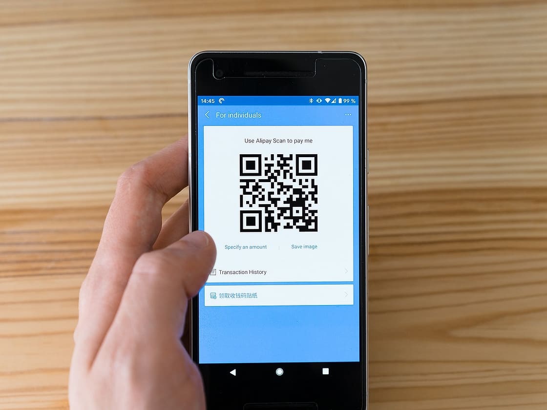 Dłoń trzyma smartfon z kodem QR do płatności nad drewnianym blatem